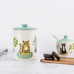 Op de afbeelding: Keramische pot met een teal deksel en een ontwerp van een gele en oranje kat die in een groene plantenbak met groene planten zit. Op de achtergrond staat een kleinere teal keramische kom met een zwart kattenontwerp en een lepel.