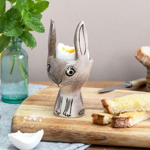 May include: A ceramic egg cup shaped like a rabbit with a speckled grey finish, painted facial features, and long ears. The cup holds a soft-boiled egg with a visible yellow yolk. The rabbit design includes black line details for eyes, whiskers, and paws.
