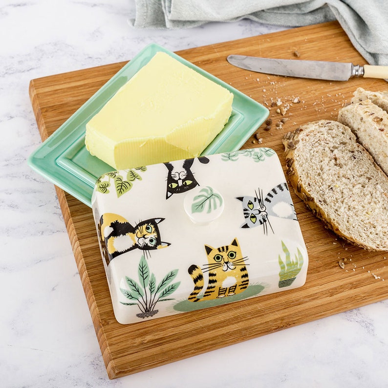 Puede incluir: Un mantequillero de cer&aacute;mica con fondo blanco y un dise&ntilde;o de gato colorido. La tapa tiene un pomo de hoja verde. El mantequillero est&aacute; lleno de un bloque de mantequilla amarilla.