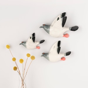Peut inclure: Trois sculptures murales d'oiseaux en céramique en plein vol, avec des corps blancs, des têtes grises, des ailes et des queues aux pointes noires, et de petites pattes roses. Les oiseaux sont disposés en diagonale, avec un vase en verre transparent.