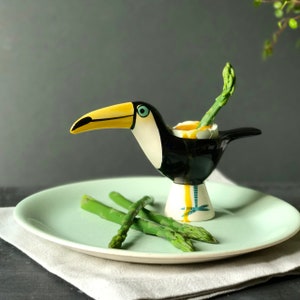 May include: A ceramic toucan egg cup with a yellow beak, black body, and white accents. The egg cup holds a soft-boiled egg with a green asparagus spear. The cup sits on a light green plate with additional asparagus spears.