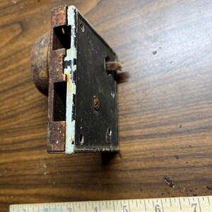 May include: An antique door lock with a rusty, round knob and a rectangular black plate. The plate has a white border with chipped paint. A wooden ruler with the text "GARDNER CONST" is in the foreground.