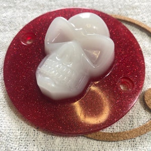 May include: A white skull-shaped resin ornament on a red glitter background. The skull has a detailed face with a white, iridescent finish.