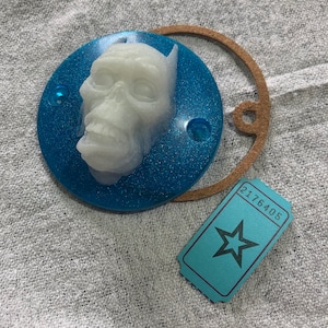 May include: A blue and white round resin ornament with a 3D sculpted white face with a toothy grin. The ornament has a glittery blue finish.