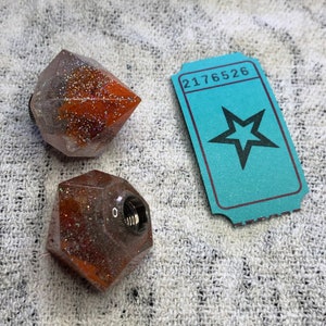 Può includere: Due tappi per bottiglia geometrici in resina trasparente con glitter arancioni e argentati. I tappi hanno un inserto metallico filettato. I tappi sono su una superficie bianca testurizzata. Un biglietto blu con il numero 2176526 e una stella nera è sullo sfondo.