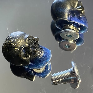 May include: Two black and blue skull-shaped drawer pulls with silver screws. The skulls have a glittery finish.