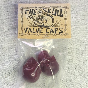 Peut inclure: Deux bouchons de valve en forme de crâne de couleur bordeaux dans un sac en plastique transparent avec une étiquette en papier brun qui indique "The Skull Valve Caps".