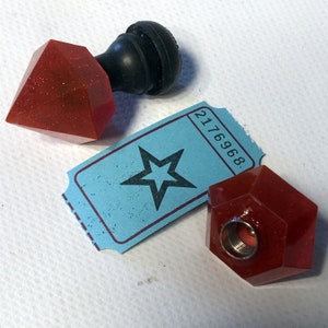 Puede incluir: Dos tapones de botella geométricos rojos, brillantes y facetados. Un tapón tiene forma de diamante y tiene un pomo negro en la parte superior. El otro tapón tiene forma de hexágono. Un billete azul con una estrella y el número 2176968 está en el fondo.