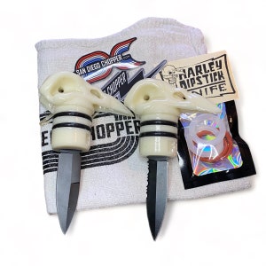 May include: Two white skull-shaped knife handles with black accents and black blades. The knives are on a white towel with a black and white logo that says "San Diego Chopper". There is a small bag with a sticker that says "Harley Dipstick Knife".