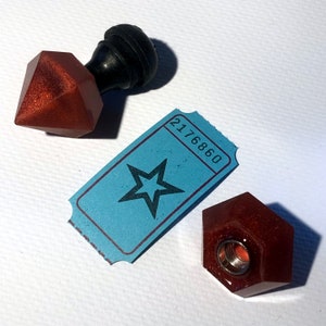 May include: Two red, sparkly, geometric bottle stoppers with a black knob on top. A blue ticket with a star and the number 2176860 is in the center of the image.