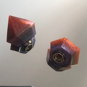 Peut inclure: Deux bouchons de bouteille en résine iridescente violet et marron en forme de diamants géométriques. Les bouchons ont un insert en métal doré.