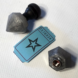 May include: Two gray metal bottle stoppers with a star-shaped ticket with the number 2176934 printed on it. One bottle stopper has a black wooden knob on top.