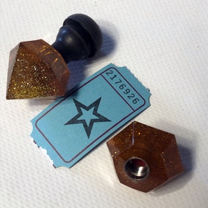 May include: Two gold glitter resin bottle stoppers, one shaped like a diamond and the other a hexagon, with a black wooden knob on top of the diamond stopper. A blue ticket with a star and the number 2176926 is also in the image.