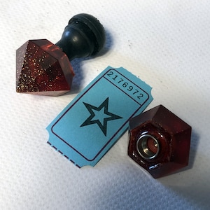 Peut inclure: Deux bouchons de bouteille en verre rouge avec une finition pailletée dorée. Un bouchon est en forme de diamant et a un bouton noir sur le dessus. L'autre bouchon est en forme d'hexagone. Un billet bleu avec le numéro 2176972 et une étoile noire est en arrière-plan.