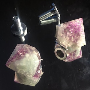 Peut inclure: Deux boutons en cristal transparent avec des paillettes violettes incrustées dans la résine. Les boutons sont fixés à des bases métalliques avec des vis.