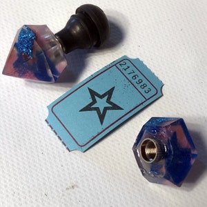 Puede incluir: Dos tapones de botella de resina con un diseño de remolino de purpurina azul y rosa. Un tapón tiene forma de diamante y el otro es un hexágono. Ambos tapones tienen un roscado de metal en la parte inferior. También hay un billete azul con una estrella y el número 2176983 en la imagen.