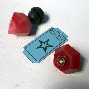 May include: Two red glass bottle stoppers with a star-shaped ticket in the background. One stopper is a diamond shape and the other is a hexagonal shape.