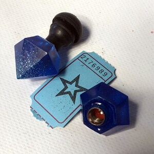Può includere: Due tappi per bottiglia in vetro blu, uno a forma di diamante con glitter e l'altro di forma geometrica, con un tappo per bottiglia nero sopra un biglietto blu con il numero 2176989.