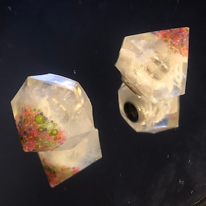 Peut inclure: Deux dés en résine transparente de forme géométrique. Les dés ont un fond blanc pailleté avec des paillettes roses, vertes et bleues incrustées à l'intérieur.