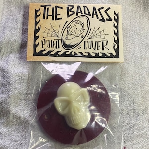 Puede incluir: Una tapa de punto en forma de calavera blanca sobre un fondo burdeos. La tapa está empaquetada en una bolsa de plástico con una tarjeta que dice "The Badass Point Cover".