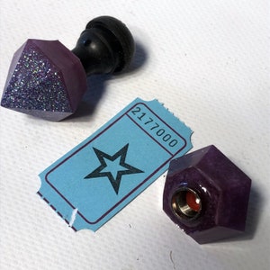 May include: Two purple glass bottle stoppers with a glittery finish. One stopper has a black knob on top. The other stopper has a gold threaded opening. A blue ticket with a star and the number 2177000 is in the background.