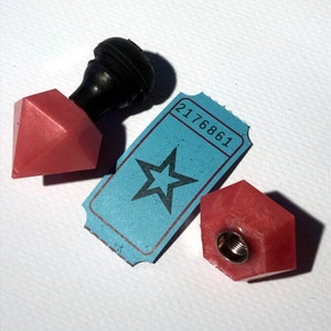 Puede incluir: Dos tapones de botella en forma de estrella rosa con un tapón de botella negro en el fondo. También se ve un billete azul con el número 2176861.