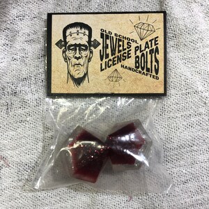 Puede incluir: Dos tornillos para matrícula de coche de color rojo y plata con purpurina, empaquetados en una bolsa de plástico transparente. La bolsa tiene una etiqueta que dice "Old School Jewels Plate License Bolts Handcrafted".