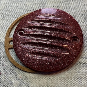 May include: A round, burgundy-colored cover with a glittery finish and horizontal grooves. Two holes are visible on the cover. A brown, circular gasket is partially visible next to the cover. The items are on a gray, textured surface.