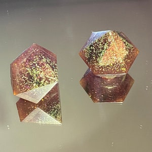 Peut inclure: Deux formes géométriques en résine iridescente de couleur violette et dorée avec des paillettes. Les formes sont toutes les deux multi-facettes et ont un aspect moucheté.