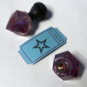 May include: Two purple glass bottle stoppers with a star-shaped ticket with the number 2176978. The stoppers have a faceted design and a threaded opening.