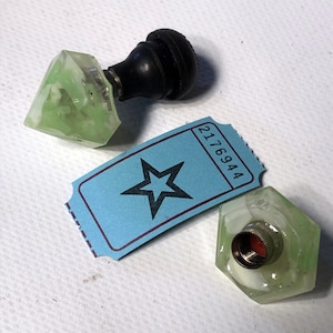 May include: Two green glass bottle stoppers with a black knob on top. One stopper is shaped like a diamond and the other is a hexagon. The stoppers are sitting on a blue ticket with a star and the number 2176944.
