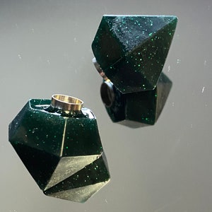 Peut inclure: Deux bouchons de bouteille en résine de couleur vert foncé, de forme géométrique et avec une finition scintillante. Un bouchon a un anneau en métal argenté autour du haut.