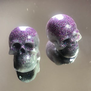 May include: Two small, white resin skull figurines with purple glitter accents.