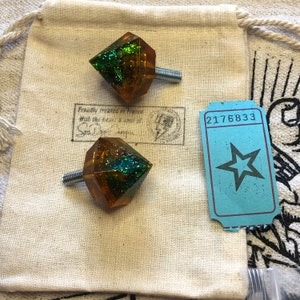 May include: Two green and gold diamond-shaped drawer pulls with silver screws. The pulls are sitting on a white fabric surface with a ticket that says "2176833" on it.