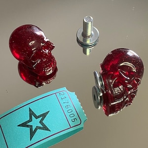Peut inclure: Deux tirettes de tiroir en forme de crâne en verre rouge avec des vis et des rondelles en métal argenté. Les vis sont fixées aux crânes. Un billet bleu avec une étoile et le numéro 2176005 est au premier plan.