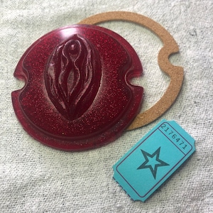 Könnte beinhalten: Eine rote, glitzernde, runde Kunststofflinse mit einem erhabenen Design. Die Linse hat zwei Ausschnitte an der Seite. Eine braune, runde Pappeinlage befindet sich neben der Linse. Ein blaues Ticket mit einem Stern und der Nummer 2176471 befindet sich in der unteren rechten Ecke des Bildes.