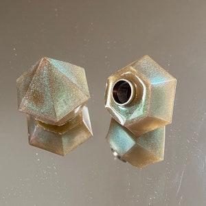 Può includere: Due tappi per bottiglia geometrici iridescenti. I tappi hanno un design sfaccettato e una lucentezza metallica con sfumature di verde e blu. Un tappo ha un inserto metallico filettato.