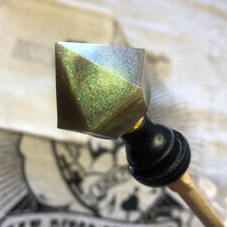 May include: A close-up of a decorative, iridescent, diamond-shaped bottle stopper with a black and gold base. The stopper has a glittery, green and gold finish.