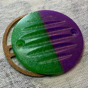 May include: A two-toned, green and purple, circular object with a textured surface and two holes. The object is next to a brown, circular gasket. The green side has a glittery effect.