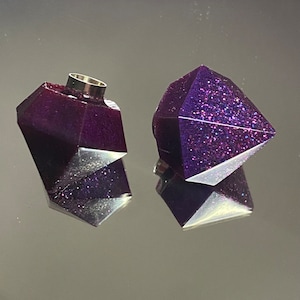 Puede incluir: Dos tapones de botella en forma de diamante de resina púrpura con purpurina. Un tapón tiene un inserto de metal plateado.