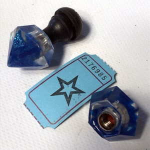 May include: Two blue resin bottle stoppers with a black knob on top. The stoppers are shaped like a diamond and a hexagon. A blue ticket with a black star and the number 2176985 is in the background.