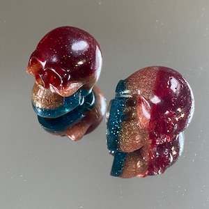 Peut inclure: Deux petites décorations de crâne en résine avec une finition pailletée rouge, or et bleu.