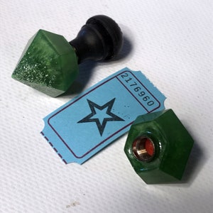 May include: Two green glass bottle stoppers with a star-shaped ticket in the background. One stopper is a diamond shape with a black knob on top. The other stopper is a hexagonal shape with a gold-colored screw top.