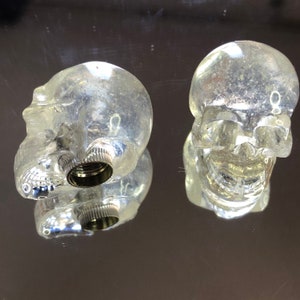 Peut inclure: Deux bouchons de bouteille en plastique transparent en forme de crâne avec une base filetée argentée.