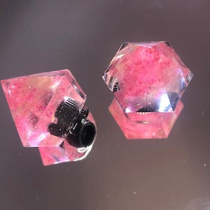 Puede incluir: Dos tapones de botella de resina rosa con purpurina. Uno tiene forma de diamante y el otro de hexágono.