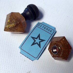 May include: Two gold glitter resin bottle stoppers shaped like diamonds with a black knob on top. One stopper has a metal screw on the bottom. A blue ticket with the number 2176906 and a black star is in the background.