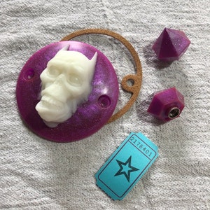 May include: A purple round resin coaster with a white skull design. The coaster is decorated with glitter. There are two purple resin bottle stoppers and a blue ticket with the number 2176401 printed on it.