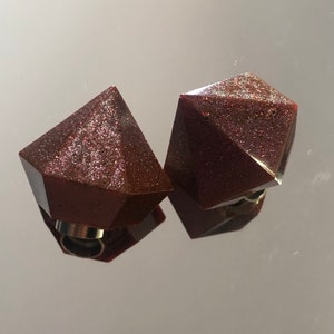 Puede incluir: Dos colgantes geométricos de resina de color rojo oscuro y brillante con bucles de metal plateado.