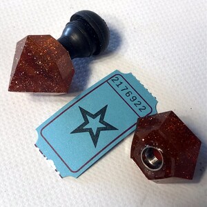 Peut inclure: Deux bouchons de bouteille en paillettes brunes avec un bouton noir sur le dessus. Un bouchon a un ticket avec le numéro 2176922 et une étoile imprimée dessus.