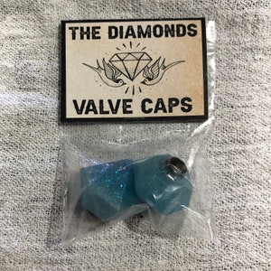 Può includere: Due tappi per valvole blu con glitter e un anello argentato in cima. I tappi sono confezionati in un sacchetto di plastica trasparente con un cartoncino bianco su cui è scritto "THE DIAMONDS VALVE CAPS".